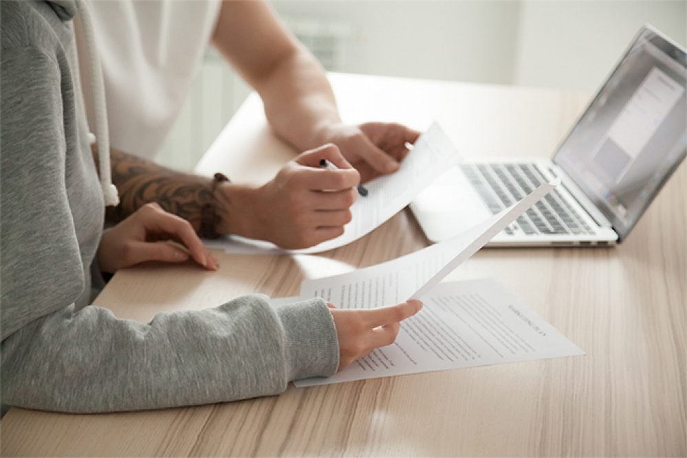 Couple reading legal documents at home with laptop, family considering mortgage loan or insurance, studying contract details, discussing terms and conditions, close up view of hands holding papers